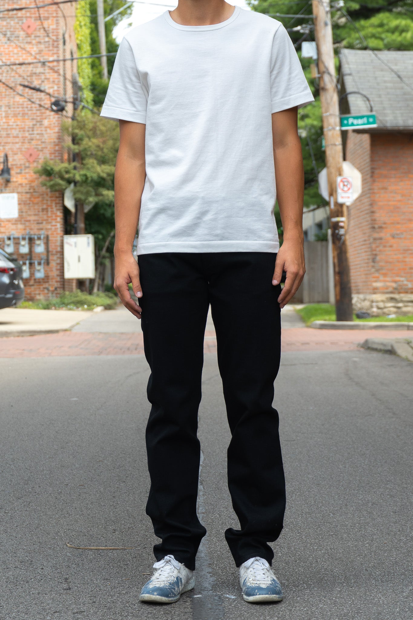 A person standing on a city street wearing a plain white short-sleeve crewneck T-shirt, black jeans, and white sneakers