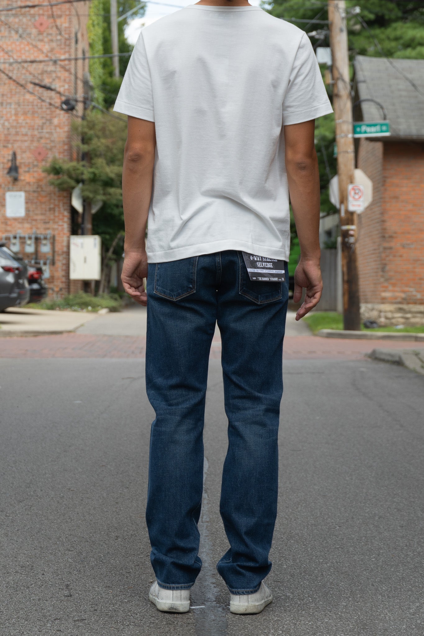 A person standing on a street, viewed from behind, wearing a white t-shirt and medium wash blue jeans. T