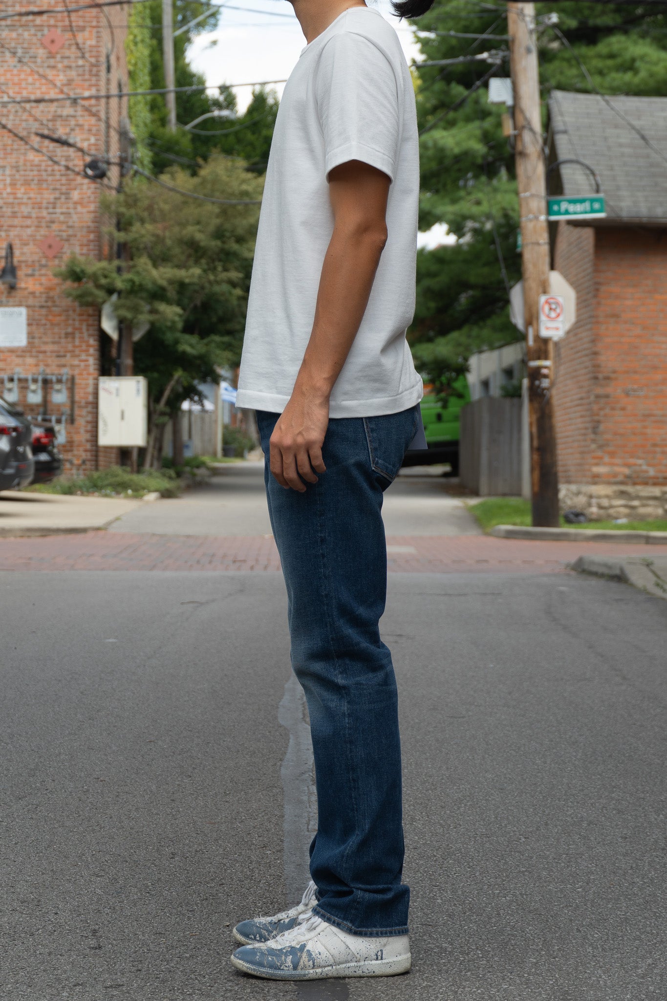 A person standing outdoors on a street wearing a plain white short-sleeve T-shirt, medium blue jeans, and white sneakers