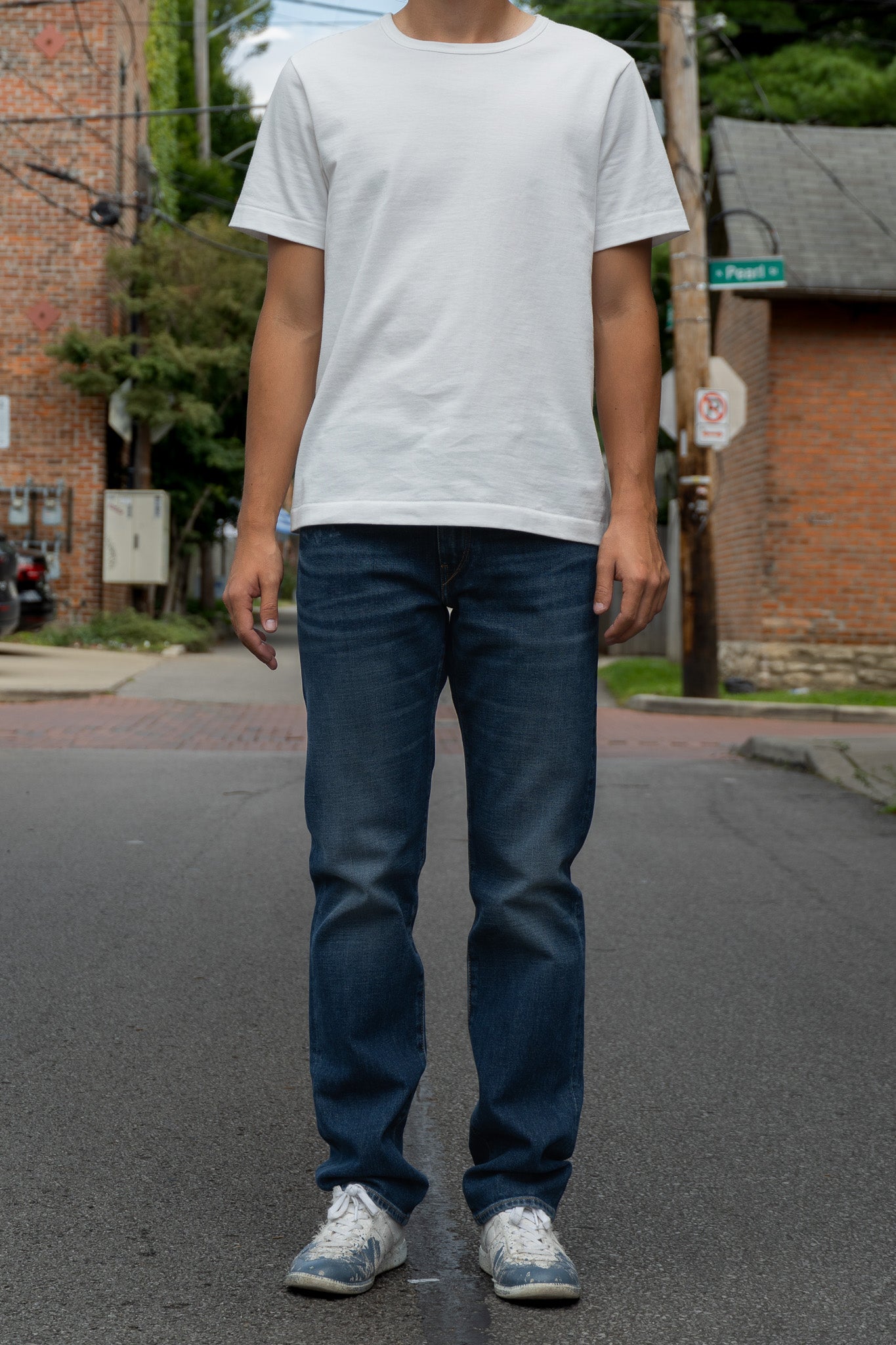 A person standing on a street wearing a plain white short-sleeve t-shirt, medium wash blue jeans, and white sneakers.