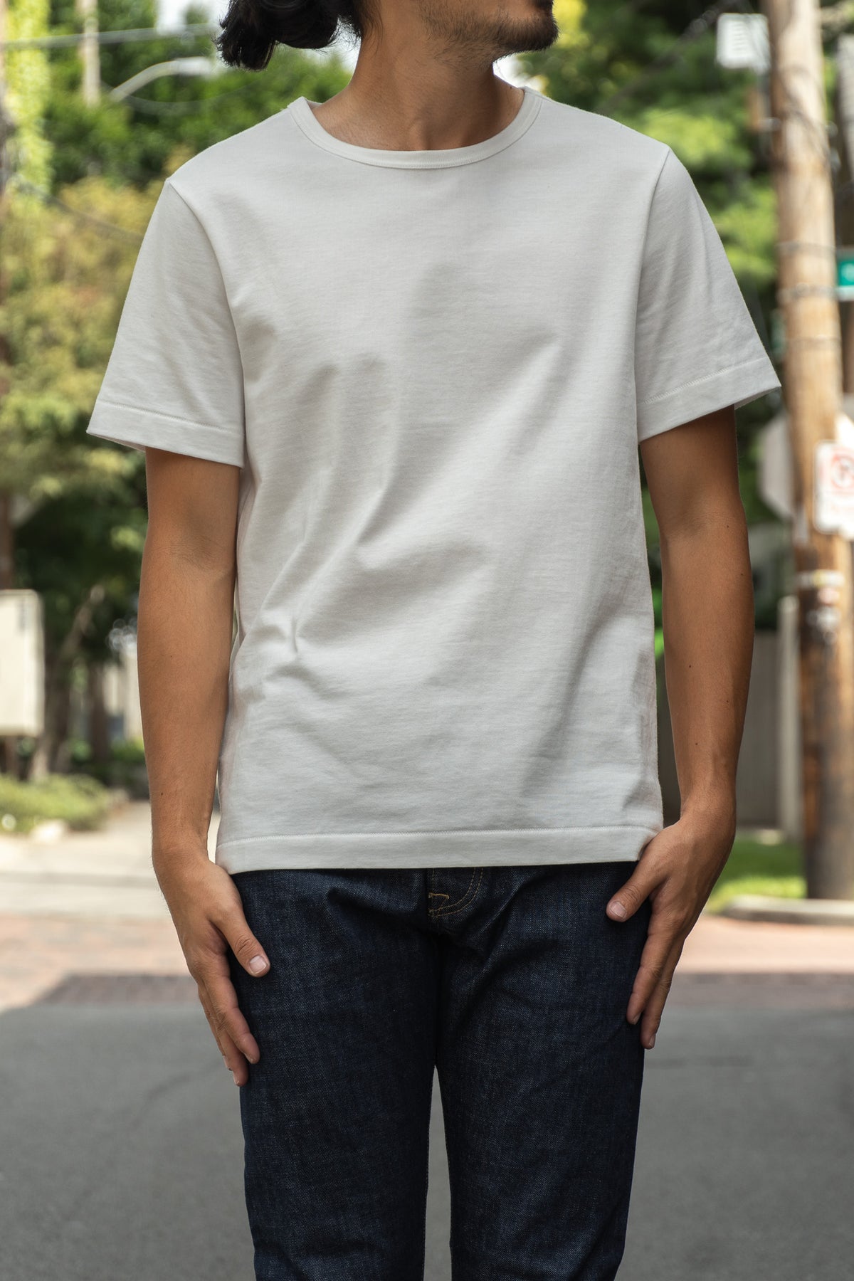 A man wearing a plain white crew-neck t-shirt and dark blue jeans, standing outdoors on a street 