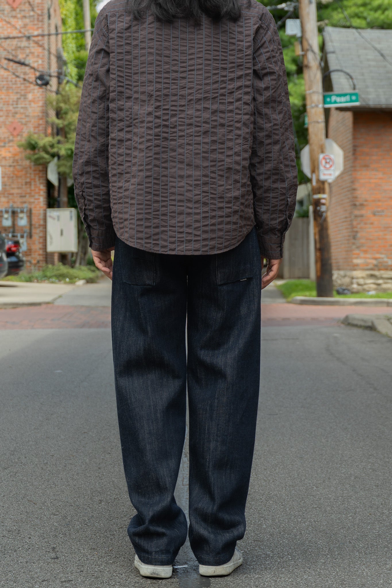 A person standing on a city street, viewed from behind, wearing a brown seersucker shirt and dark blue jeans.