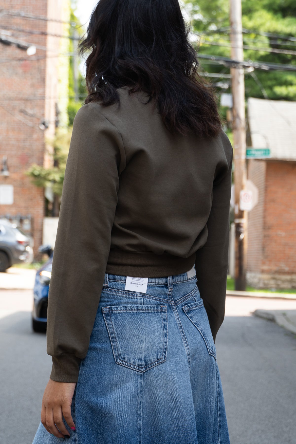 A woman viewed from behind wearing an olive brown long-sleeve cropped zip up and a high-waisted blue denim skirt in an outdoor street setting.