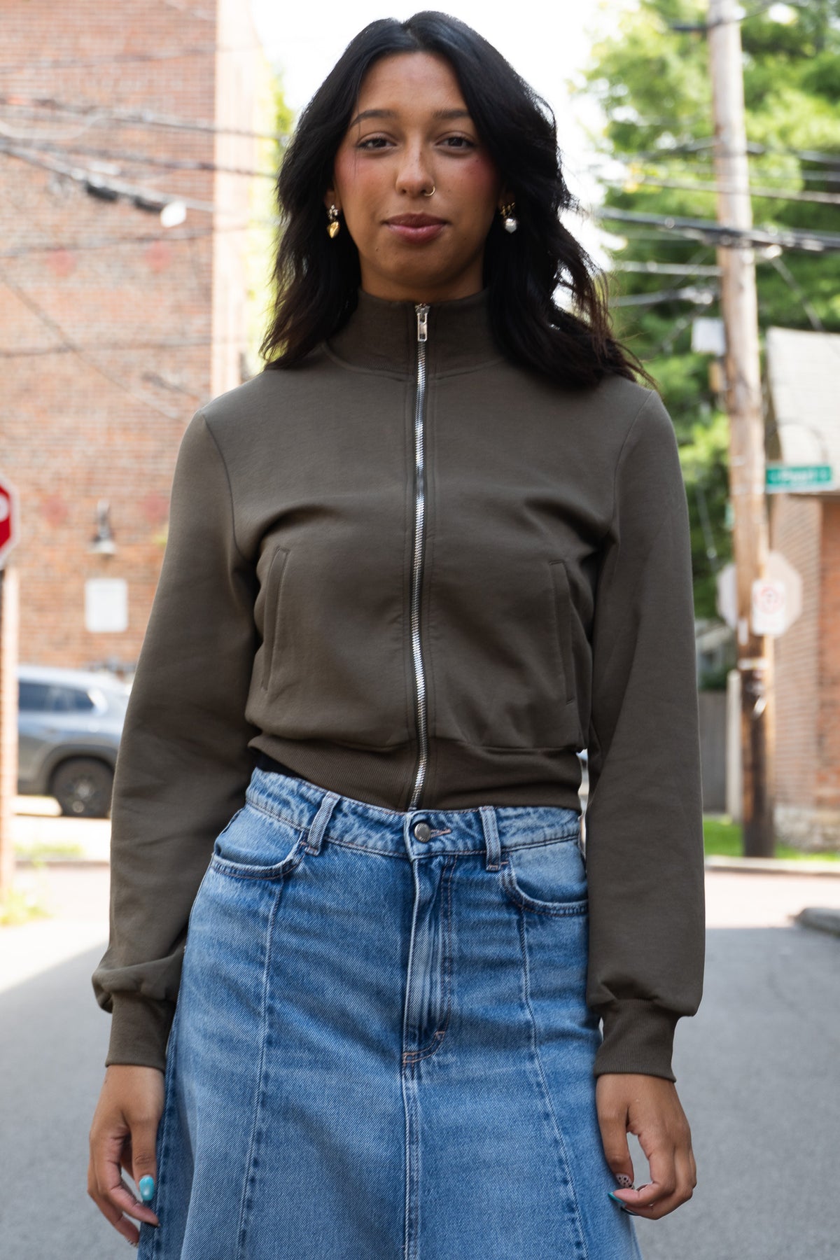 A woman wearing a fitted olive-brown zip-front jacket with a mock neck and ribbed cuffs, paired with a blue denim skirt, standing in the street