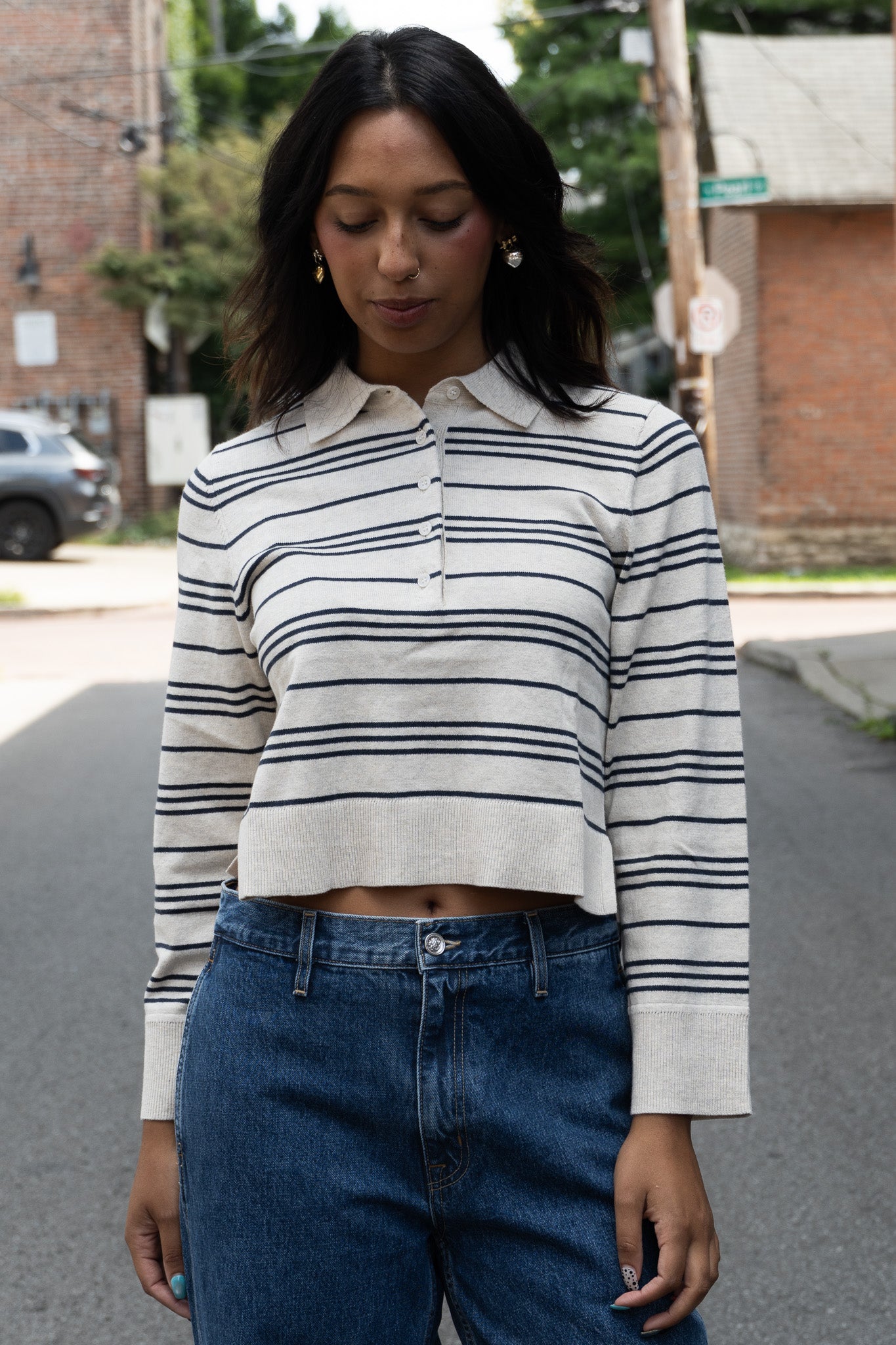 A woman wearing a cream and navy blue horizontally striped long-sleeve cropped polo shirt with dark blue jeans, standing on an outdoor street.