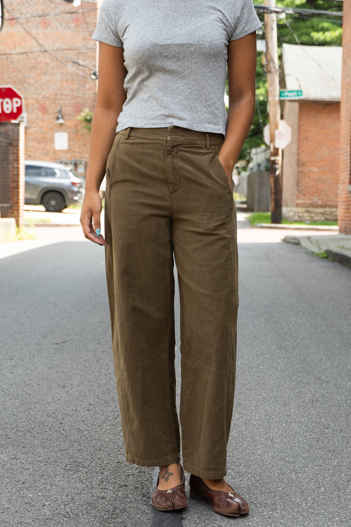 A person standing on an outdoor street wearing high-waisted, wide-leg olive pants, a light gray t-shirt, and brown leather loafers.