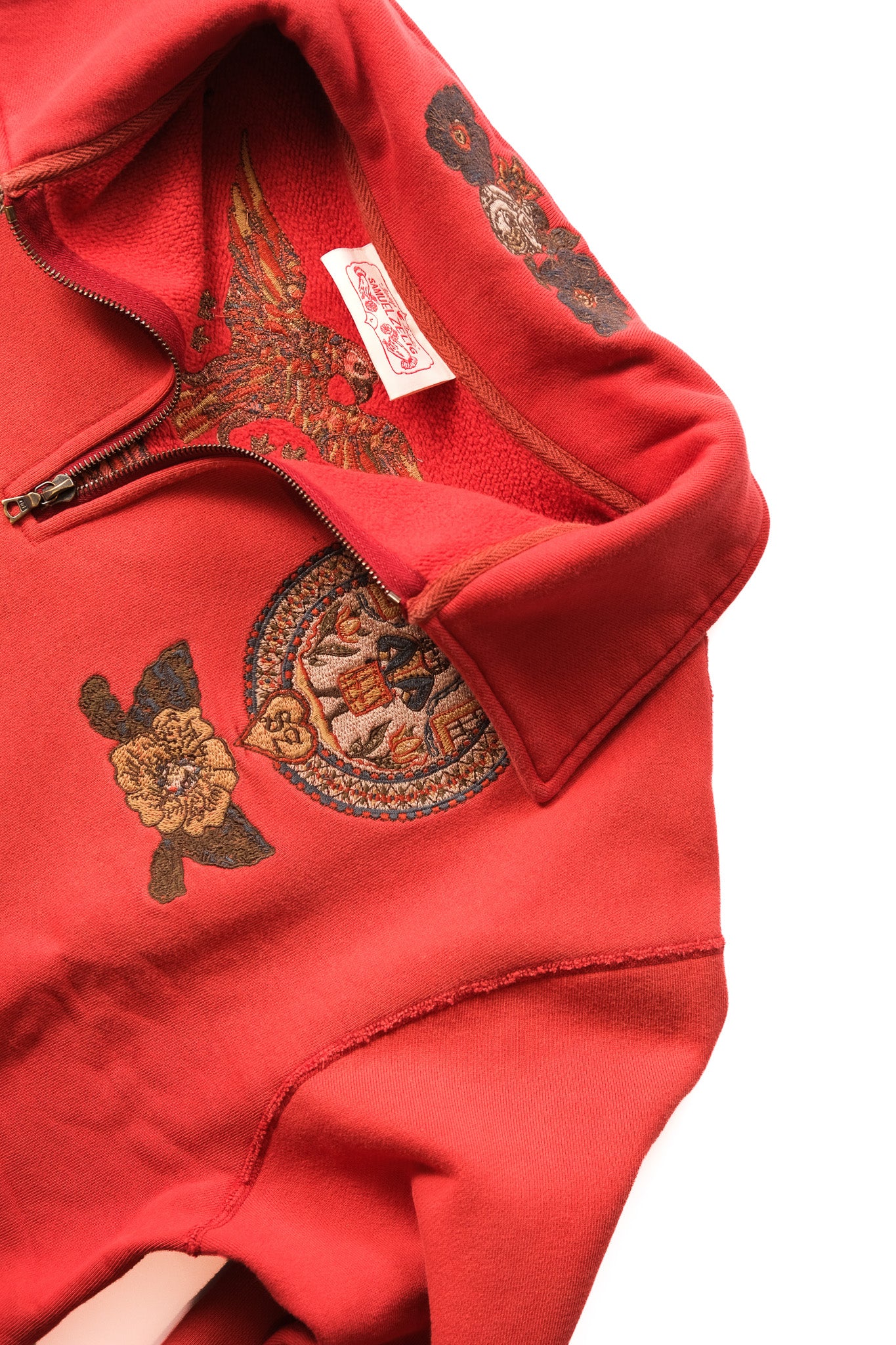 A close-up of a red fleece pullover 1/4 zip featuring embroidered details, including a circular medallion and floral motifs on the chest, with a white brand tag visible inside the collar.