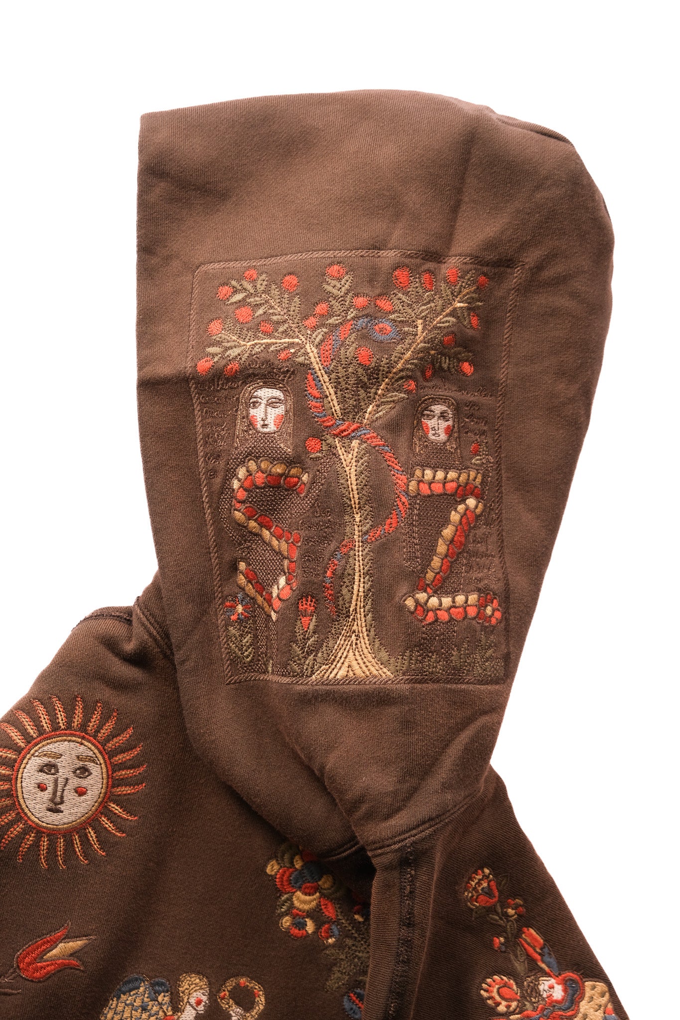 A close-up of a brown hooded garment featuring intricate embroidered folk-art motifs, including a tree with red berries, coiled snakes, sun faces, and small figures, shown against a plain white background.