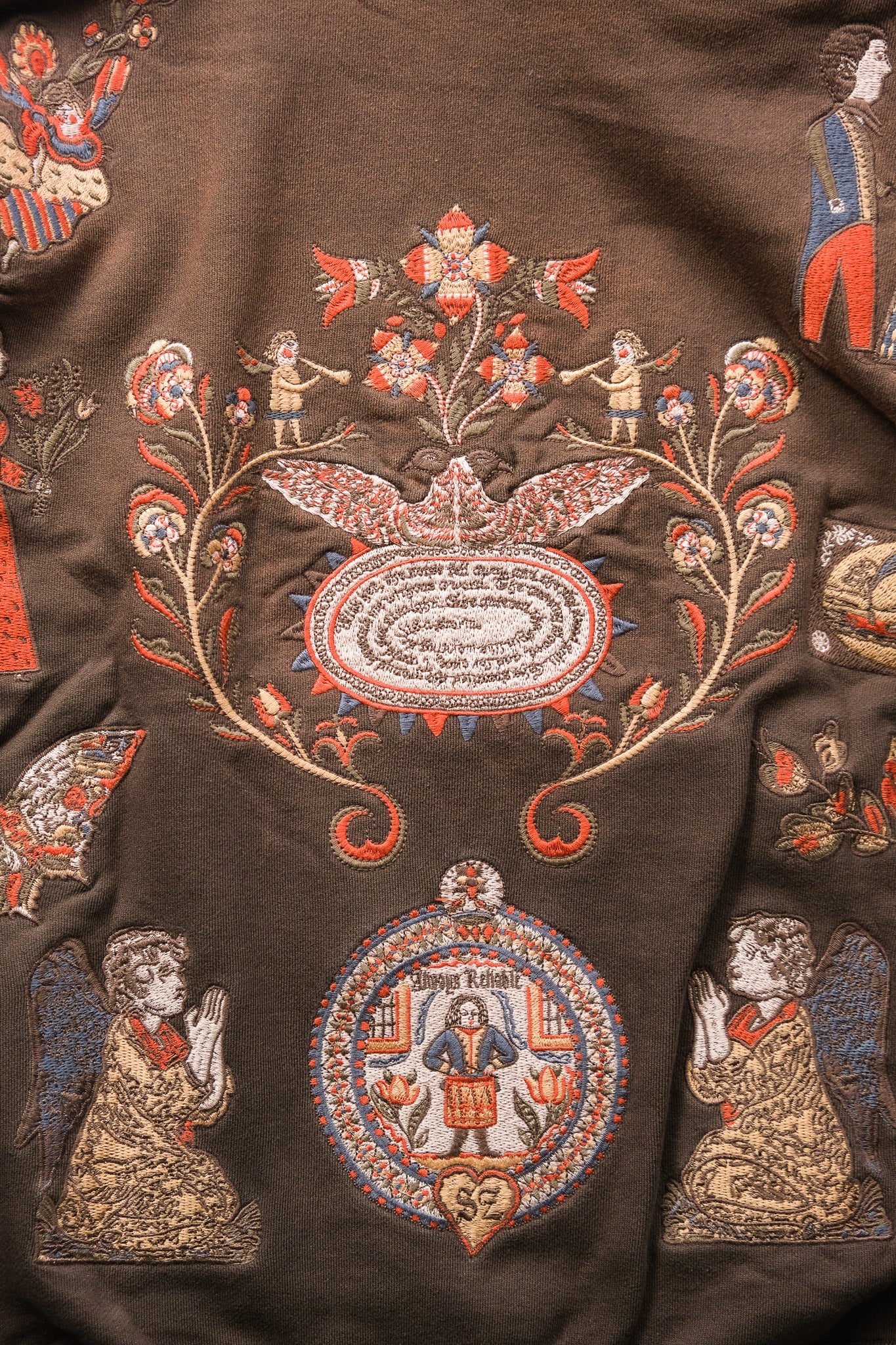 A close-up of a dark brown embroidered textile featuring ornate floral motifs, angel figures, and circular medallions in red, beige, and blue thread.