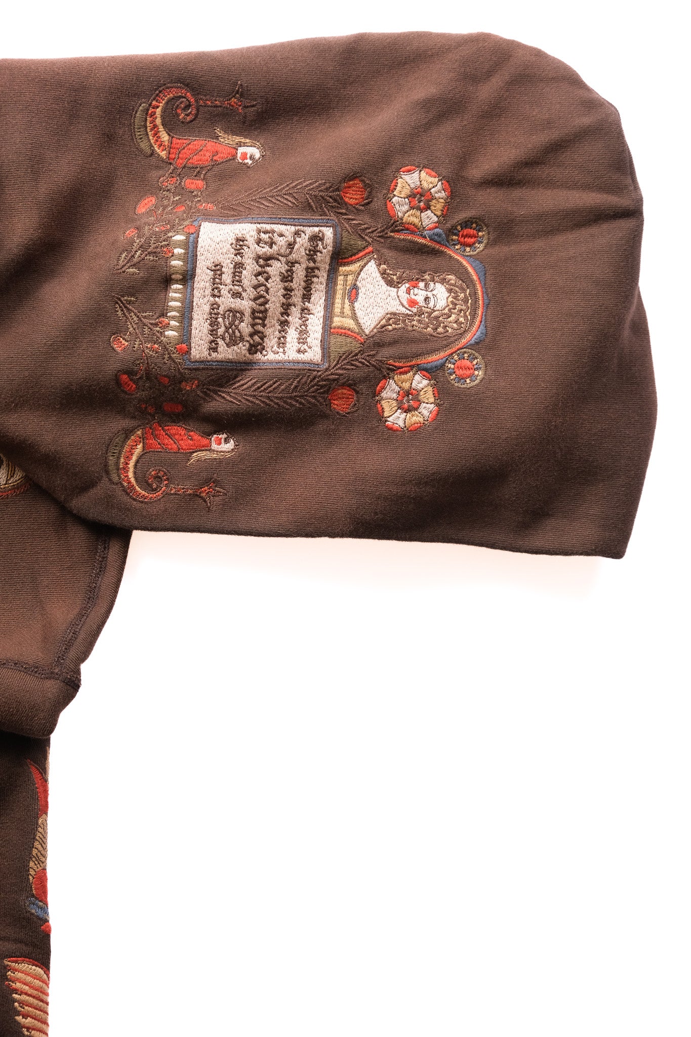 A close-up of a brown garment featuring a detailed folk embroidery with a central label, set against a plain white background.
