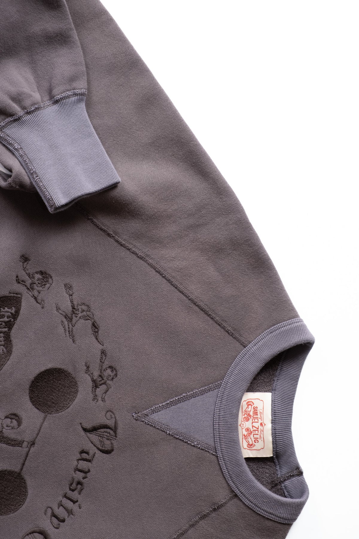 Close-up of a dark gray sweatshirt showing the sleeve cuff, ribbed collar, and a tonal embroidered graphic print on the front, with a small red-and-white brand tag visible inside the collar.