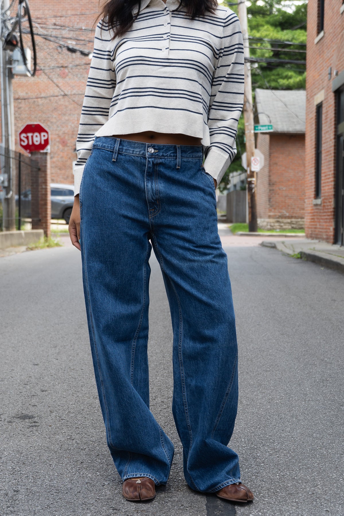 A person standing on a street wearing blue wide-leg jeans, a cream and navy striped long-sleeve cropped top, and brown shoes.